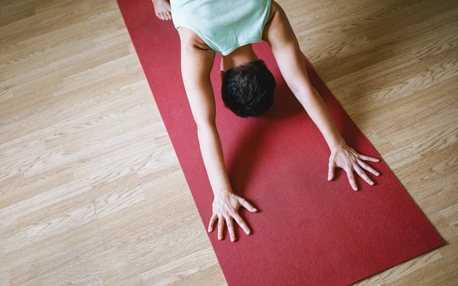 Seen from above, a person lowers into a stretch on a red mat on a wooden floor